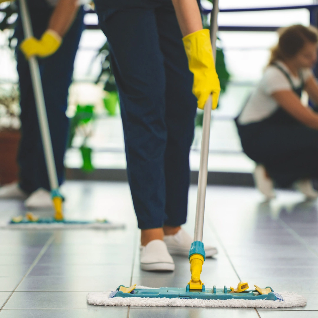 A person in yellow gloves uses a flat mop to clean a tiled floor, with other members of a cleaning crew visible in the background.