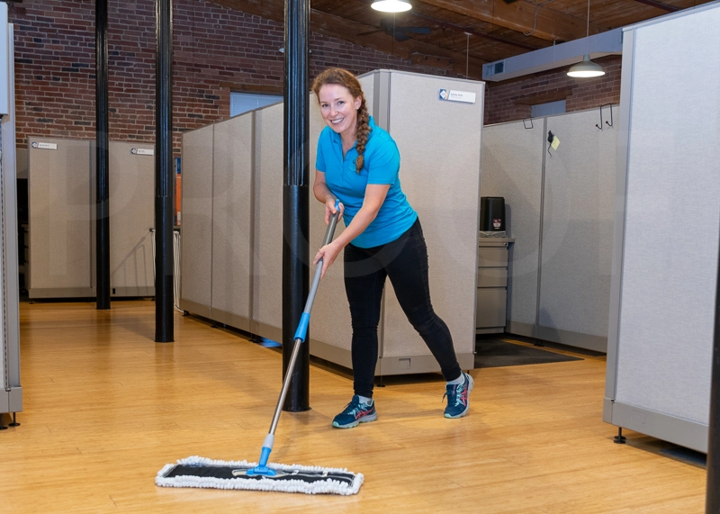 A person in a blue shirt mops the floor of a modern office space with cubicles and exposed brick walls