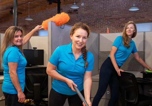 Three women in matching blue polo shirts pose with cleaning supplies, including a mop and an orange duster, inside an office cubicle area.